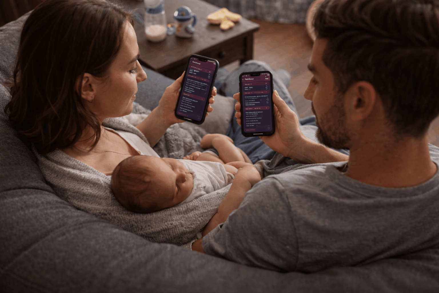 Two parents checking a synced baby tracker app on their phones while caring for their baby